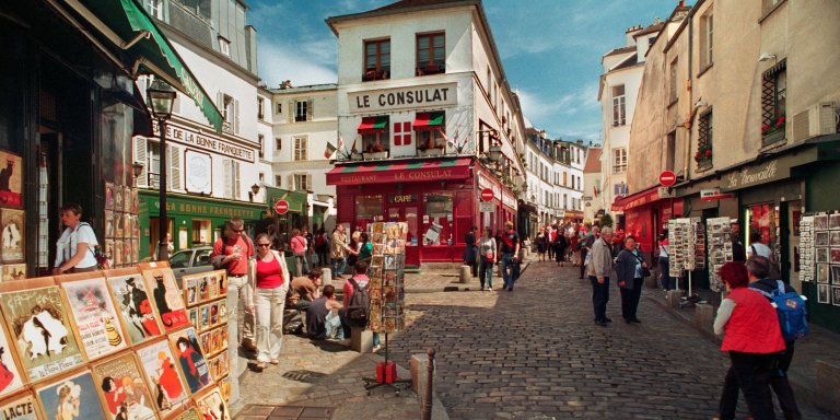 street view in montmartres with cobble stones, tourist walking down the street, painting on the left side, and a quaint restaurant called le Consulat in the background.