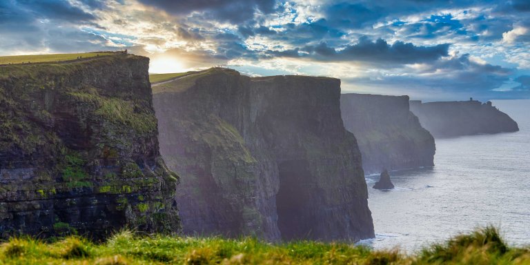landscape image of the Cliffs of Moher, with sunbeams in the background shining through a cloudy blue sky